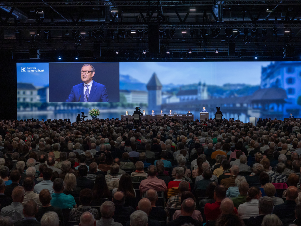 grosse Leinwand mit Rednerpult und Gästen an der GV der LUKB